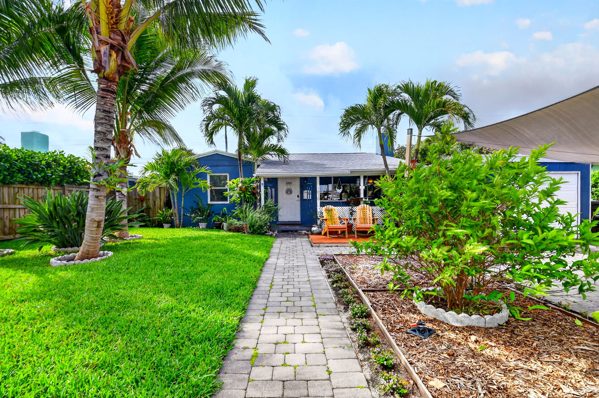 Charming blue cottage in West Palm Beach with tropical landscaping, palm trees, lush lawn, and inviting front porch.